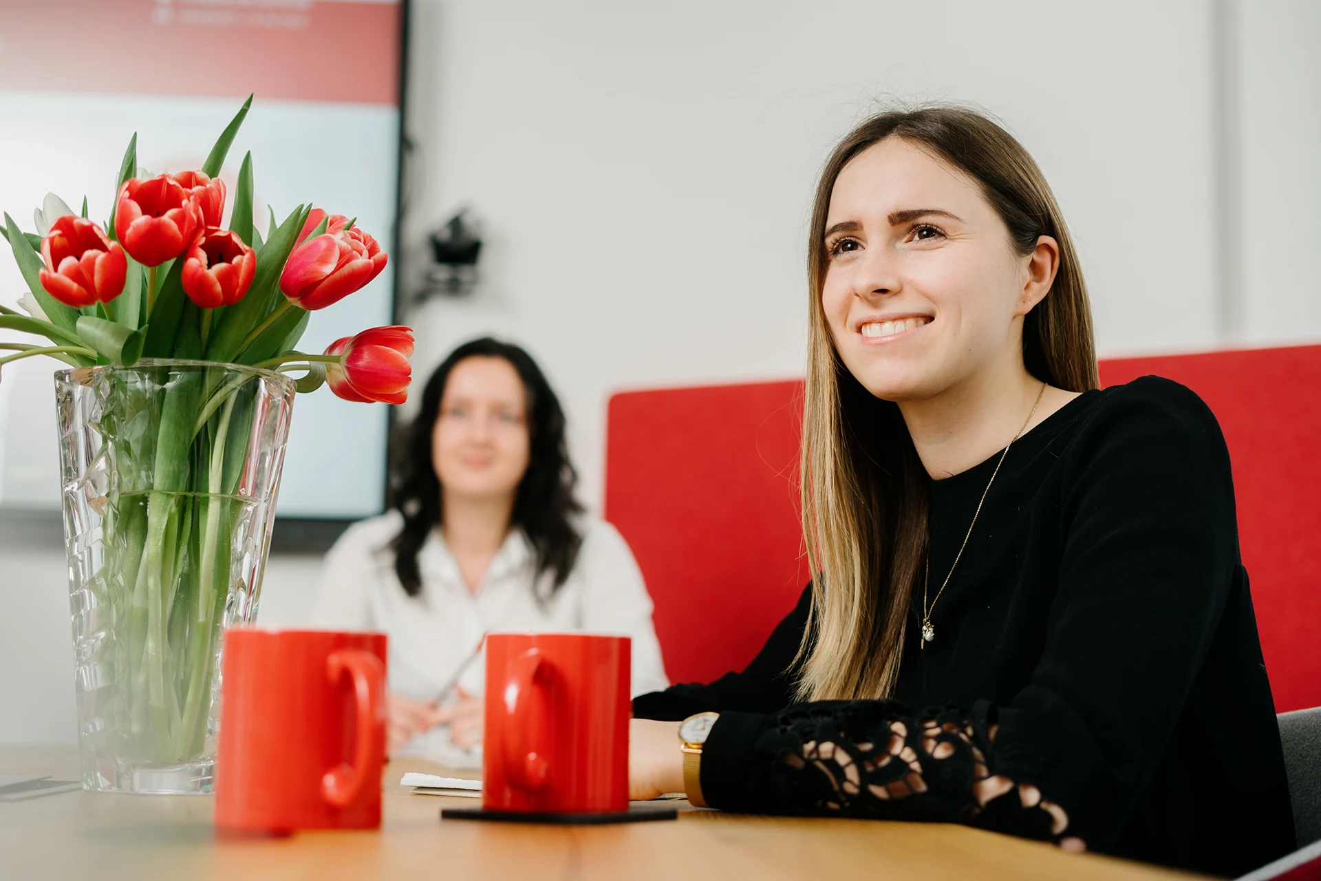 kanzlei-zeindl-partner-steuerberater-index-headerslider-01 Junge Frau lächelt während eines Gesprächs in einem modernen Besprechungsraum. Auf dem Tisch rote Kaffeetassen und ein Blumenstrauß mit roten Tulpen. Im Hintergrund eine weitere Person leicht unscharf