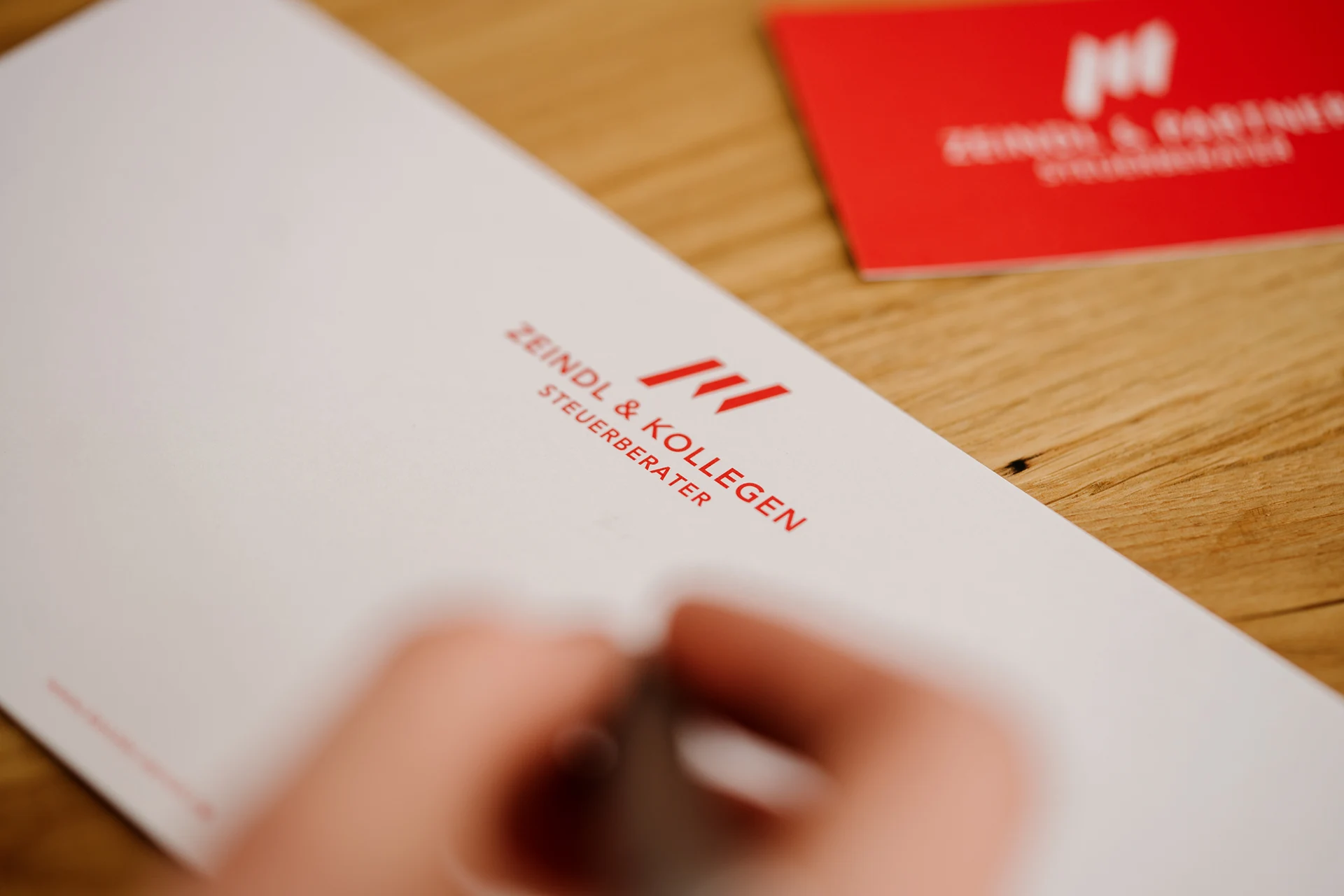 kanzlei-zeindl-partner-steuerberater-index-headerslider-03 Briefpapier mit dem roten Logo und Schriftzug ‚Zeindl & Kollegen Steuerberater‘ auf einem Holztisch, im Hintergrund unscharf eine rote Visitenkarte; im Vordergrund eine Hand, die mit einem Stift schreibt