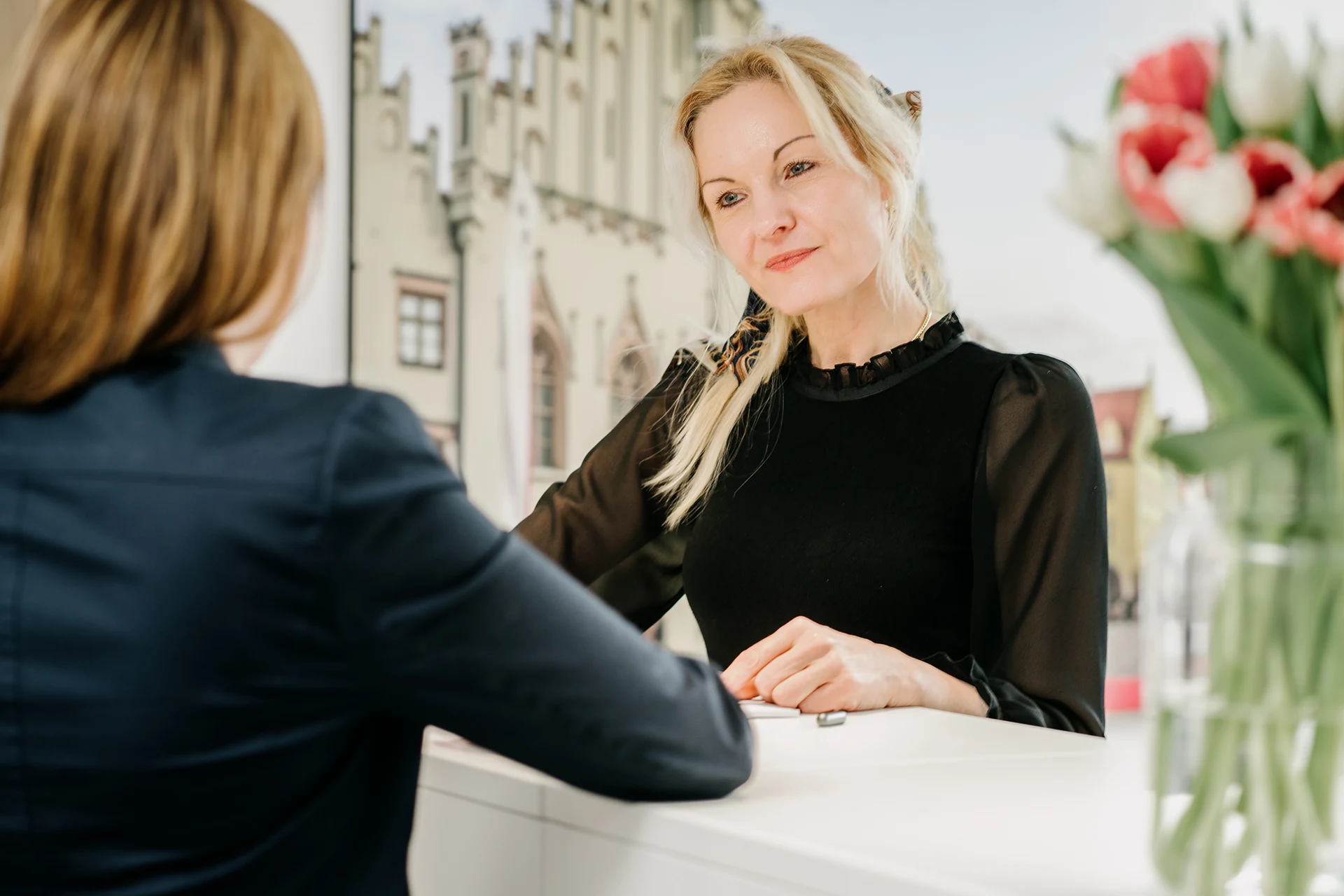 kanzlei-zeindl-partner-steuerberater-index-headerslider-04 Zwei Frauen im Gespräch an einem Empfangstresen, eine Frau blickt freundlich und aufmerksam auf ihr Gegenüber