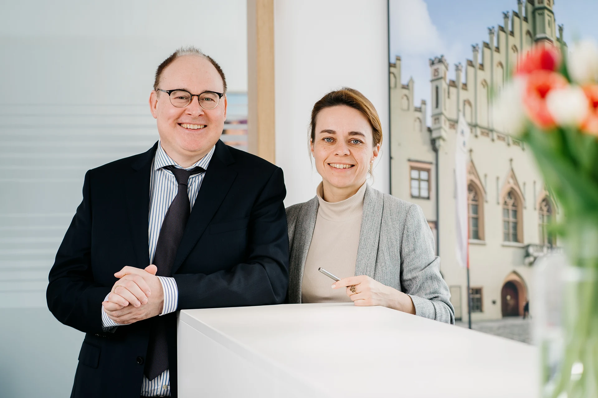 kanzlei-zeindl-partner-steuerberater-index-headerslider-05 Ein Mann im Anzug und eine Frau im grauen Blazer stehen lächelnd an einem weißen Empfangstresen, im Hintergrund ein Bild eines historischen Gebäudes