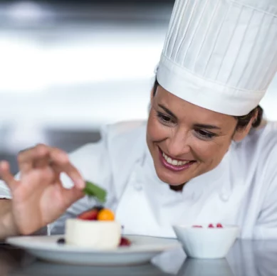 Köchin in weißer Uniform und Mütze dekoriert lächelnd ein Dessert auf einem Teller mit frischen Beeren und einem Minzblatt