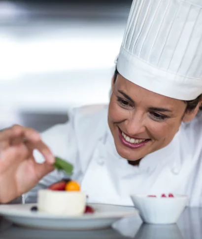 kanzlei-zeindl-partner-steuerberater-branchen-gastronomie-systemgastronomie-hotellerie Köchin in weißer Uniform und Mütze dekoriert lächelnd ein Dessert auf einem Teller mit frischen Beeren und einem Minzblatt