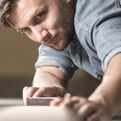 Ein Handwerker in Jeanshemd schleift konzentriert ein Werkstück. In der Brusttasche steckt ein Stift