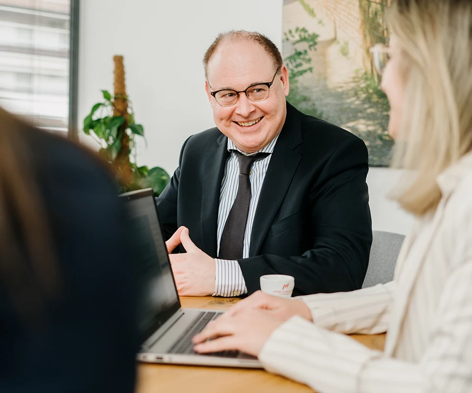 Ein Mann im Anzug und eine Frau im hellen Blazer sitzen gemeinsam an einem Tisch und unterhalten sich freundlich. Vor ihnen stehe ein geöffneter Laptop