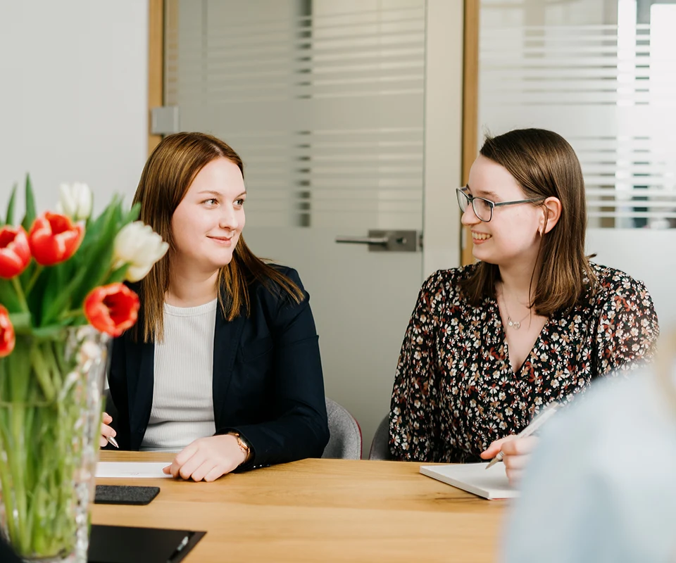 Zwei junge Frauen sitzen lächelnd an einem Besprechungstisch mit Notizblöcken. Auf dem Tisch ein Blumenstrauß mit roten und weißen Tulpen