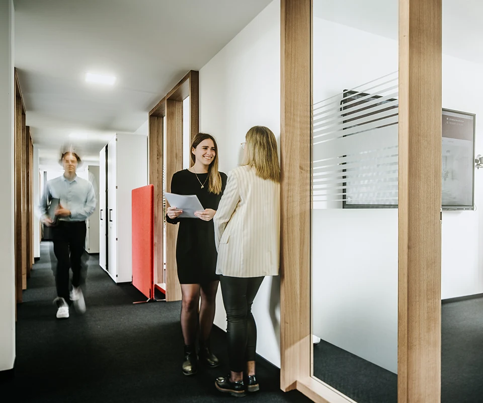 Zwei Frauen unterhalten sich lächelnd im Flur eines Bürogebäudes, links geht ein Mann dynamisch durch den Flur