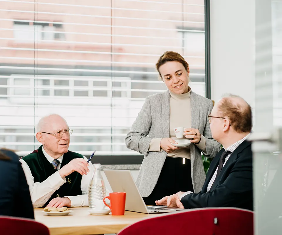 Drei Personen in Businesskleidung unterhalten sich freundlich in einem modernen Büro. Ein älterer Mann und ein Mann mittleren Alters sitzen an einem Tisch mit Laptops und Kaffee, eine Frau steht daneben mit einer Kaffeetasse in der Hand