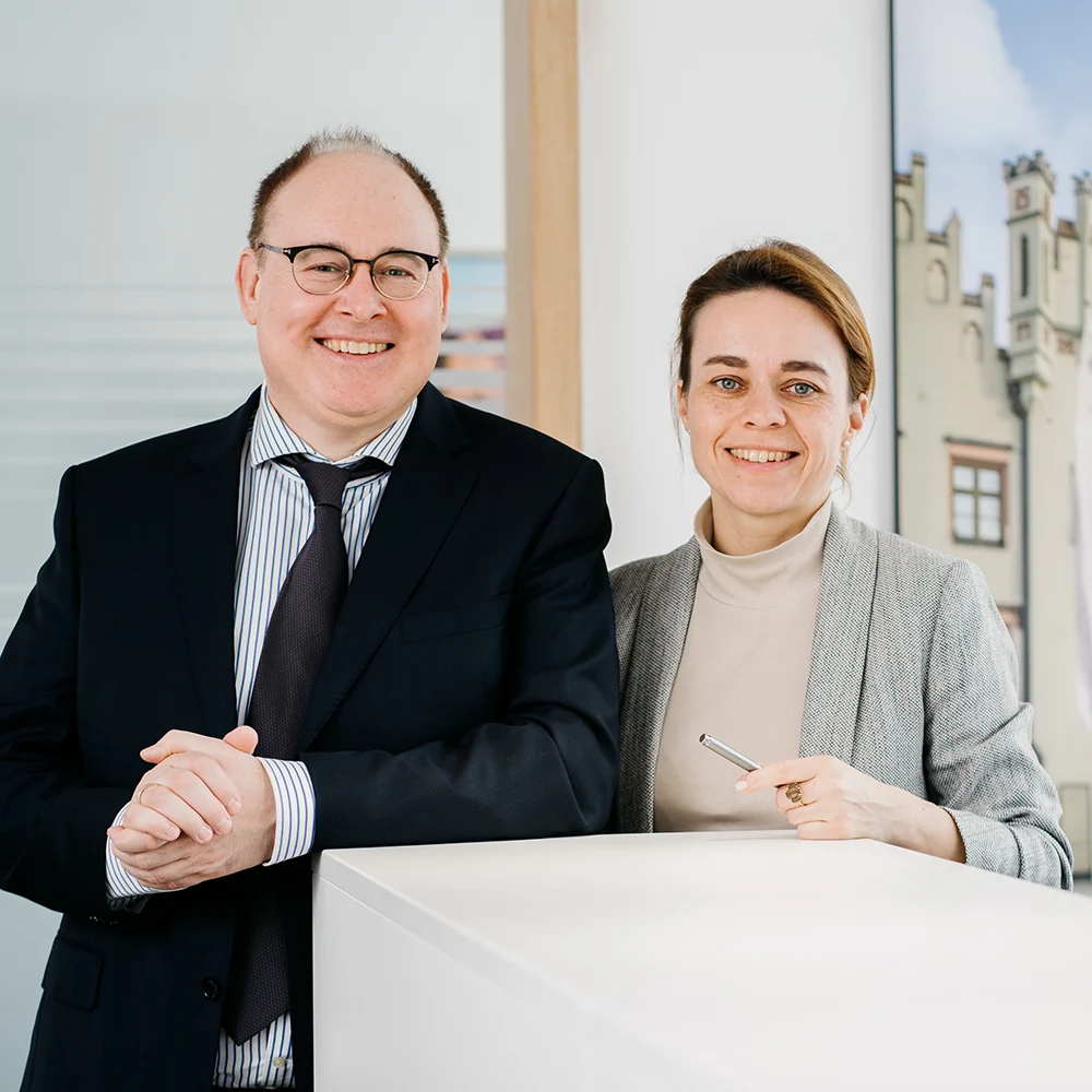 Ein Mann im Anzug und eine Frau im grauen Blazer stehen lächelnd an einem weißen Empfangstresen, im Hintergrund ein Bild eines historischen Gebäudes