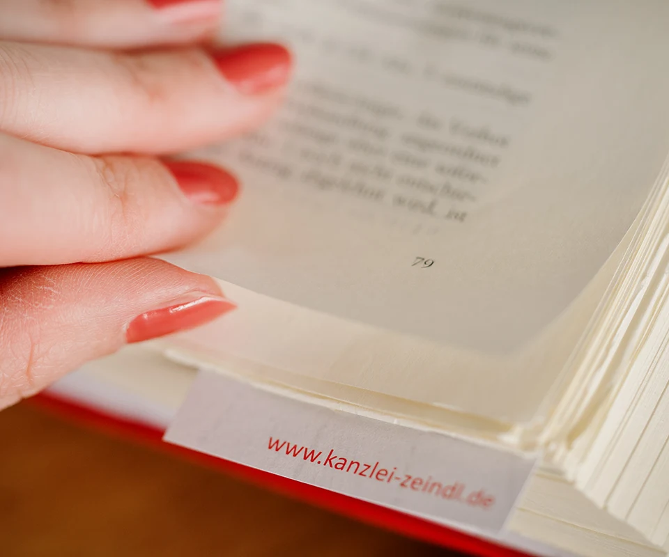 Nahaufnahme einer Hand, die eine Buchseite mit einem Registerreiter umblättert. Auf dem Reiter steht die Webadresse ‚www.kanzlei-zeindl.de‘ in roter Schrift