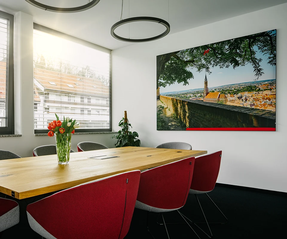 Heller Besprechungsraum mit großem Holztisch, grauen und roten Stühlen, einem Blumenstrauß mit Tulpen auf dem Tisch und einem großen Wandbild mit Stadtansicht