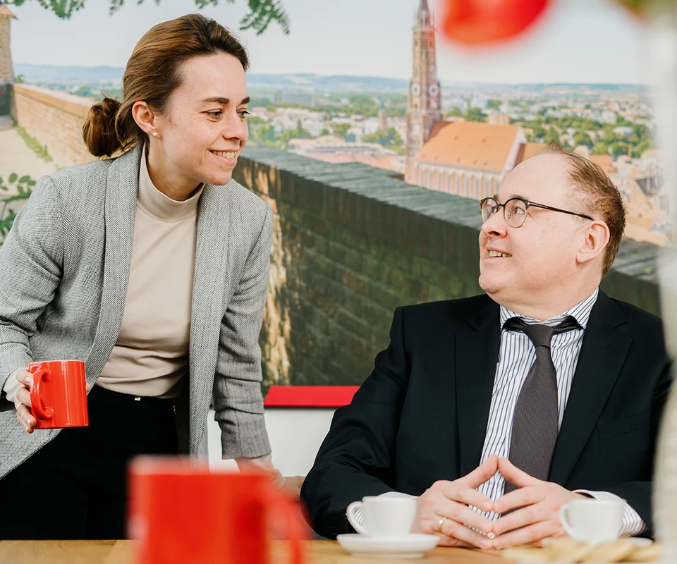 Eine Frau mit grauem Blazer und roter Tasse in der Hand beugt sich freundlich zu einem Mann im Anzug, der sie anlächelt. Im Hintergrund ist ein großes Wandbild mit der Ansicht einer historischen Stadt zu sehen