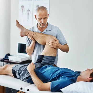 Physiotherapeut behandelt einen Patienten auf einer Behandlungsliege, der Therapeut beugt vorsichtig das Bein des liegenden Patienten