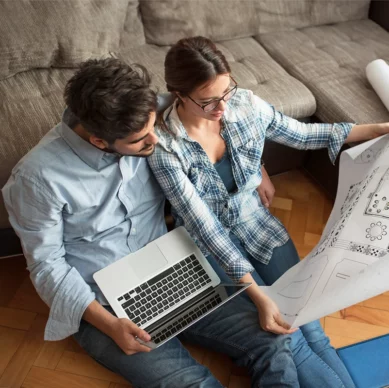 Ein junges Paar, welches auf dem Boden vor dem Sofa sitzt, die Frau hält einen Bauplan in der Hand, der Mann arbeitet an einem Laptop