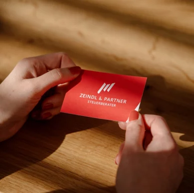 Zwei Hände halten eine rote Visitenkarte mit dem weißen Logo der Kanzlei Zeindl & Partner über einem Holztisch