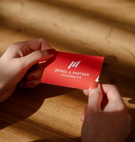 Zwei Hände halten eine rote Visitenkarte mit dem weißen Logo der Kanzlei Zeindl & Partner über einem Holztisch