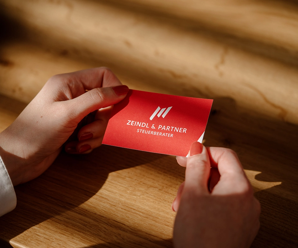Zwei Hände halten eine rote Visitenkarte mit dem weißen Logo der Kanzlei Zeindl & Partner über einem Holztisch