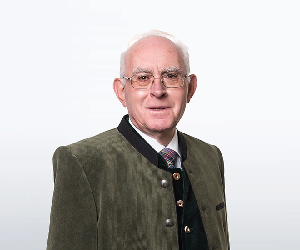 Älterer Mann mit Brille und traditioneller grüner Trachtenjacke, freundlich lächelnd vor hellem Hintergrund