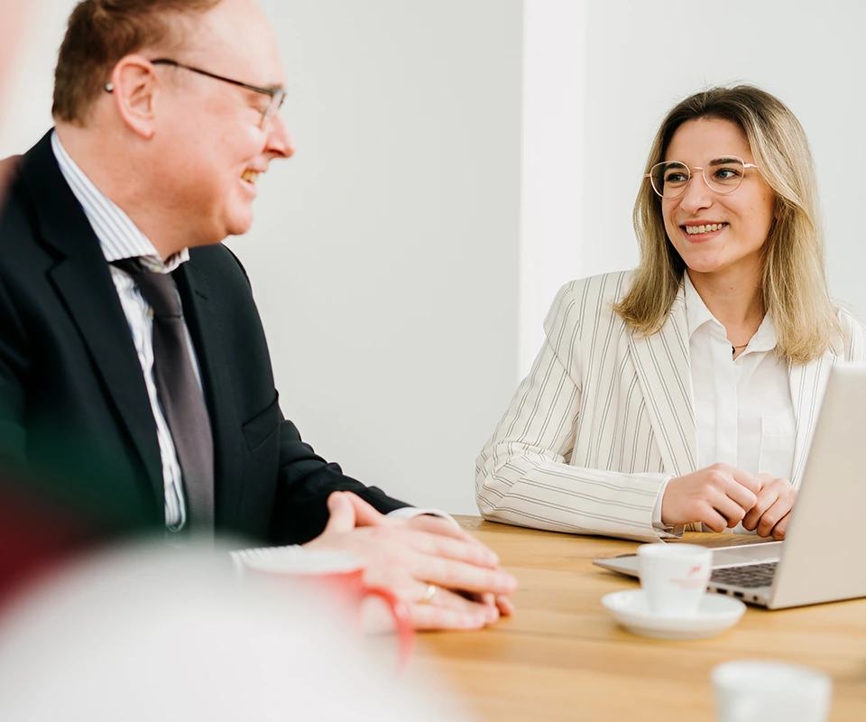 Zwei Personen in Businesskleidung sitzen an einem Tisch, unterhalten sich freundlich und lächeln. Vor ihnen ein Laptop und Kaffeetassen
