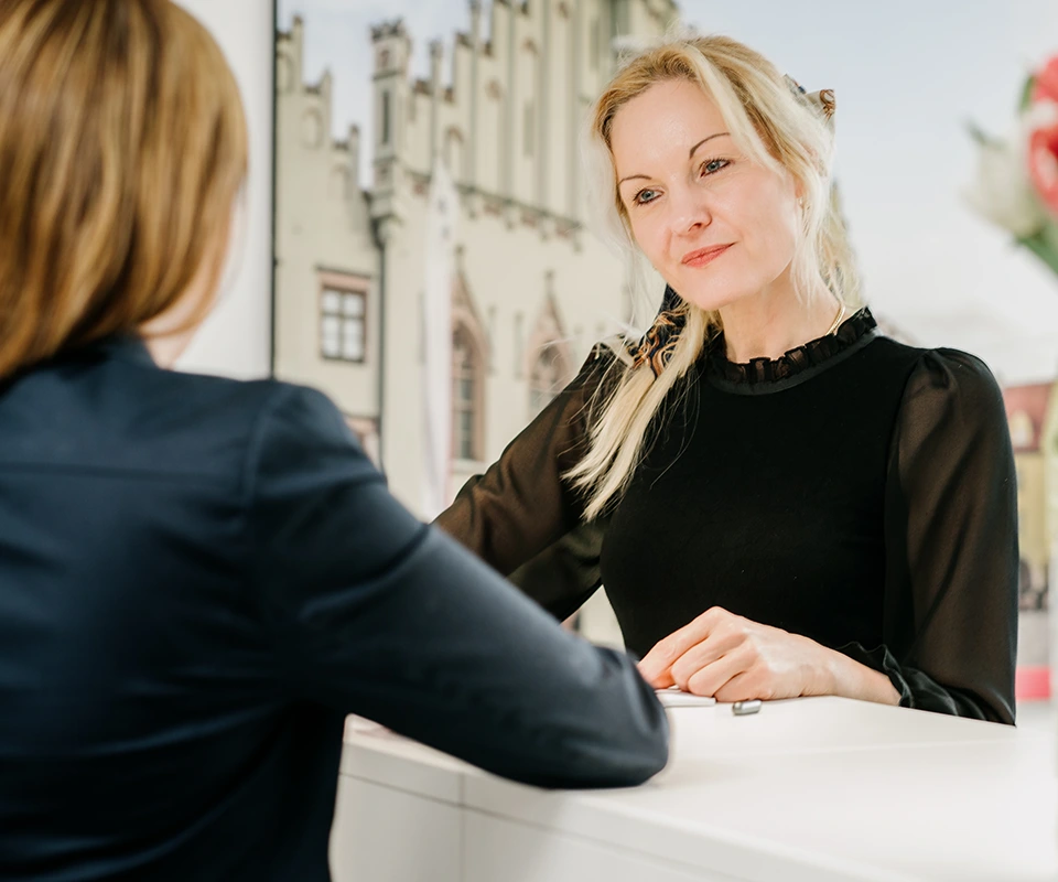 Zwei Frauen im Gespräch an einem Empfangstresen, eine Frau blickt freundlich und aufmerksam auf ihr Gegenüber